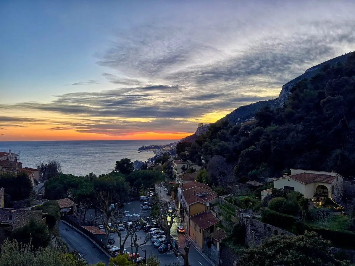 An orange sunset over the sea, seen past a wooded slope and rooftops on the hillside