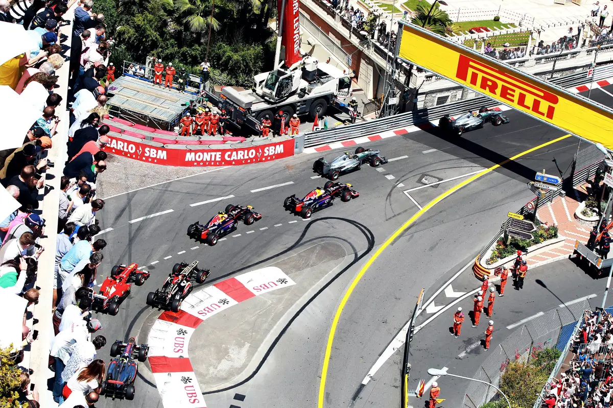 Formula One cars filing through a tight bend of the Monaco street circuit, seen from high above, with crowds behind the barriers