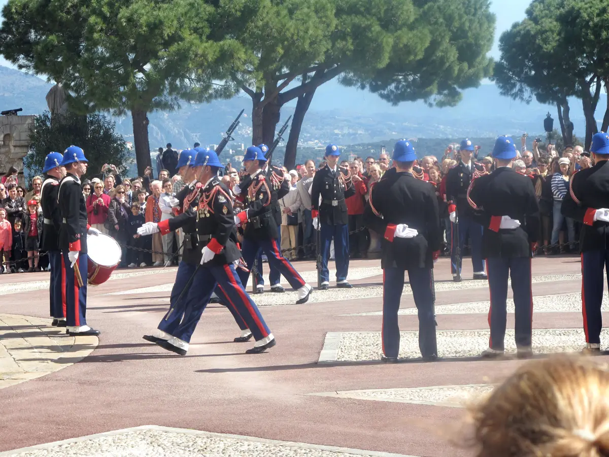 Palace guards in dark uniforms and white helmets marching past a crowd of onlookers under umbrella pines