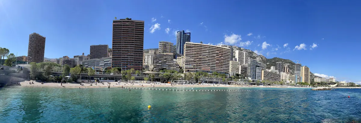 Clear turquoise water at Larvotto with the beach and the tall apartment blocks of the eastern quarters behind, under a blue sky