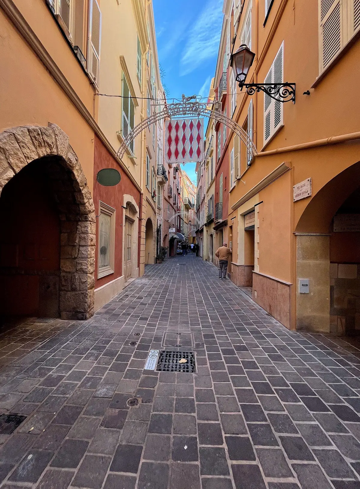 A narrow paved lane between ochre and pink houses in the old town, under an arch, with the red and white flag above